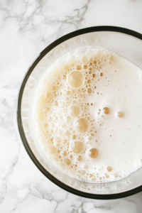 A close-up of a shiny glass mixing bowl on a white marble cooktop, filled with warm water at 110°F. A packet of active dry yeast and granulated sugar is sprinkled on top, beginning to dissolve and bubble, a crucial step in making homemade pizza dough.