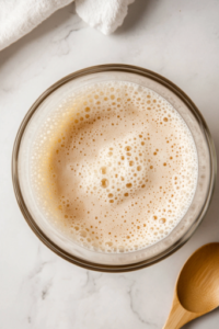 A close-up shot of a glass mixing bowl placed on a white marble cooktop, filled with warm water, sugar, and dry active yeast. The mixture appears slightly foamy, indicating that the yeast has been activated. A wooden spoon rests nearby, ready for the next step in preparing Grandma’s Perfect Homemade Bread.