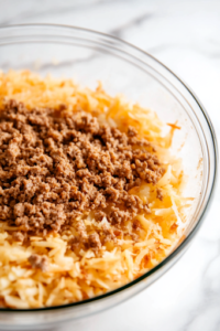 A close-up shot of the cooked ground meat being added into the large glass mixing bowl on the white marble cooktop. The browned crumbles blend seamlessly into the cheesy, seasoned hash brown mixture, creating a rich and hearty filling for the casserole.