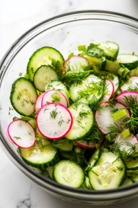 A close-up view of a large glass mixing bowl on a white marble cooktop, filled with freshly sliced cucumbers, radishes, and celery. Bright green minced dill, chives, and chopped mint are being sprinkled over the salad, adding a burst of freshness and aroma.