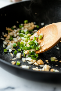 A close-up shot of the same black skillet on the white marble cooktop, with minced garlic and chopped green onions being tossed in. The garlic sizzles and turns golden, releasing a rich, fragrant aroma that enhances the stir-fry’s depth of flavor.