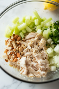A close-up shot of a glass mixing bowl on a white marble cooktop, where chopped celery, toasted almonds, mayonnaise, lemon juice, and black pepper are being added to the cooked chicken, preparing for mixing.