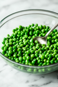A close-up shot of a large glass mixing bowl on the white marble cooktop, with freshly drained green peas being added. A spoon gently stirs them, ensuring even distribution before mixing with other ingredients.