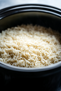 A side view of a slow cooker with uncooked long-grain white rice evenly spread on top of the cooking ingredients. Water is being poured in, allowing the rice to soak up the flavorful broth, preparing for slow and even cooking.