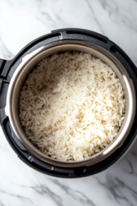 A close-up shot of an Instant Pot inner pot on the white marble cooktop, as rinsed Calrose rice is poured in, followed by 1 cup + 2 tablespoons of water. The grains glisten under the light, evenly submerged in the measured liquid.