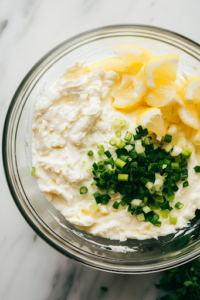 Lemon juice, Worcestershire sauce, minced garlic, and chopped green onions being added to the softened cream cheese in a glass mixing bowl on the white marble cooktop. These ingredients are being combined to enhance the flavor of the dip.