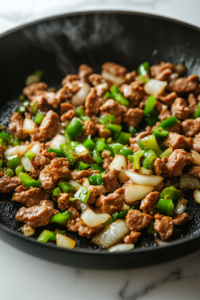 A close-up shot of the black skillet on the white marble cooktop, as minced onions, green bell peppers, and a blend of seasonings are added to the browned meat. The mixture sizzles, and aromatic steam rises, enhancing the flavors.