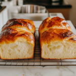A close-up of two golden brown loaves of freshly baked bread cooling on a wire rack over a white marble cooktop. The crust looks crisp, and a sliced piece reveals the soft, fluffy interior, ready to be enjoyed.