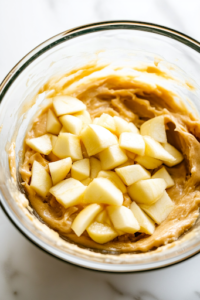 A close-up of peeled and chopped apples being gently folded into the thick cake batter inside a glass mixing bowl on a white marble cooktop. The apple pieces are evenly distributed, adding texture and flavor to the mixture.