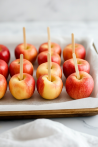 A close-up shot of a parchment-lined baking sheet placed on a white marble cooktop, lightly sprayed with cooking spray. A row of clean, dry apples stands upright with craft sticks inserted into the tops, ready to be coated with candy.