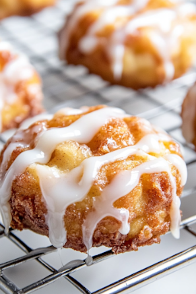 A close-up of a cooling rack on a white marble cooktop, with freshly baked apple cinnamon fritters being drizzled with smooth vanilla glaze from a spoon. The glaze cascades down, coating the warm fritters for a glossy and irresistible finish.