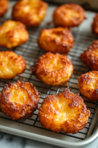 A close-up of an oven rack, showing golden-brown apple fritters baking on a parchment-lined sheet. The fritters have puffed up slightly, with crisp, caramelized edges and a warm spiced aroma filling the kitchen.