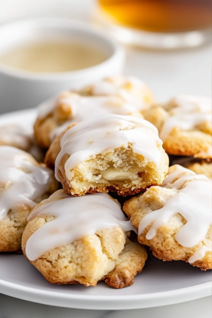 A plate of banana cookies with vanilla glaze, beautifully arranged with a bite taken from one cookie to reveal its soft, moist texture. A small dish of extra glaze and a cup of tea sit in the background.