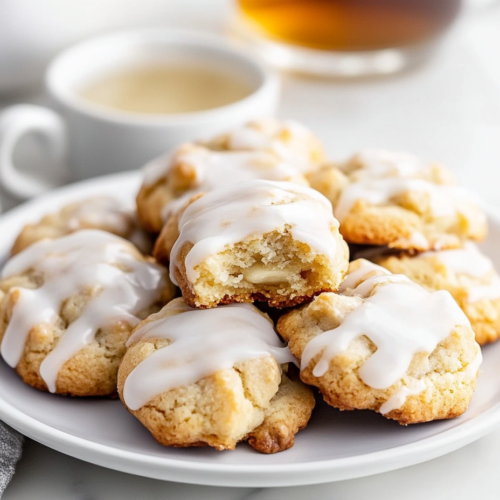 A plate of banana cookies with vanilla glaze, beautifully arranged with a bite taken from one cookie to reveal its soft, moist texture. A small dish of extra glaze and a cup of tea sit in the background.