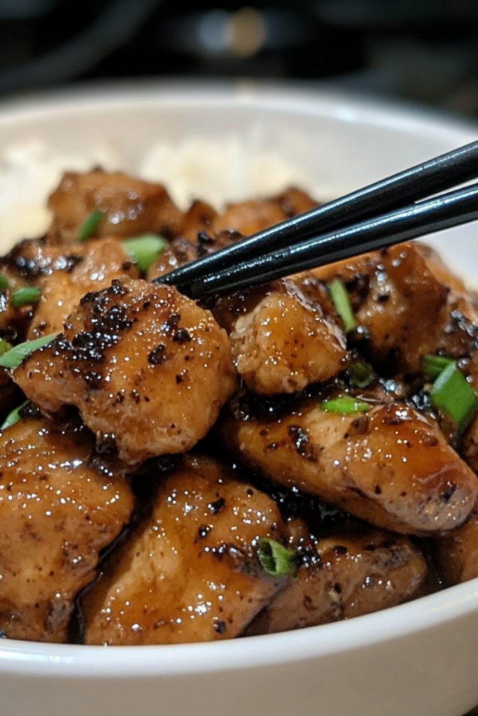 Final plated dish: a bowl of black pepper chicken stir-fry, garnished with extra black pepper, with a side of steamed white rice. A pair of chopsticks lifts a bite-sized piece of chicken, capturing its glistening, flavorful coating.