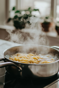 A close-up shot of a stainless steel pot on the white marble cooktop, filled with bubbling water as fettuccine pasta cooks until tender. The long pasta strands swirl in the rolling boil, while steam rises gently, signaling they are nearly ready.
