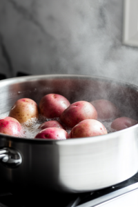 A close-up of a stainless steel pot on a white marble cooktop, filled with boiling water. Whole red potatoes are fully submerged as steam rises from the pot, indicating they are cooking until fork-tender.