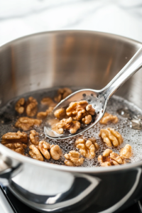 A shiny stainless steel saucepan on a white marble cooktop filled with boiling water and honey. Halved walnuts are submerged in the bubbling mixture, gently cooking to achieve a glossy, caramelized texture. A slotted spoon rests on the side, ready to remove them.