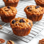 A close-up of muffins cooling on a wire rack over a white marble cooktop, with one muffin cut in half to reveal its moist, fluffy interior. A few scattered pecans and dried cherries add texture, making the muffins look even more delicious.