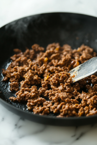 A close-up shot of a black skillet on a white marble cooktop, where ground beef and minced chicken gizzards sizzle in hot vegetable oil. A spatula breaks apart the meat as it browns, releasing a rich, savory aroma.