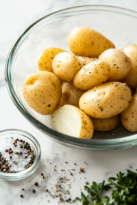 A close-up shot of a glass bowl on the white marble cooktop, where potatoes are being evenly brushed with olive oil. A small dish filled with kosher salt, black pepper, garlic powder, and dried parsley sits nearby, ready to add a flavorful seasoning.