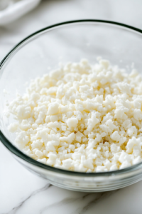 A close-up view of the same glass mixing bowl on a white marble cooktop as softened butter is added. The flour mixture takes on a coarse, crumbly texture, preparing the dough for the next step.