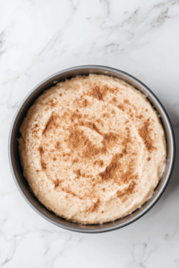 A close-up of a greased 9-inch cake pan on a white marble cooktop, with half of the cake batter being spread evenly. The cinnamon-sugar mixture is generously sprinkled on top before the remaining batter is added, creating a delicious layered effect.