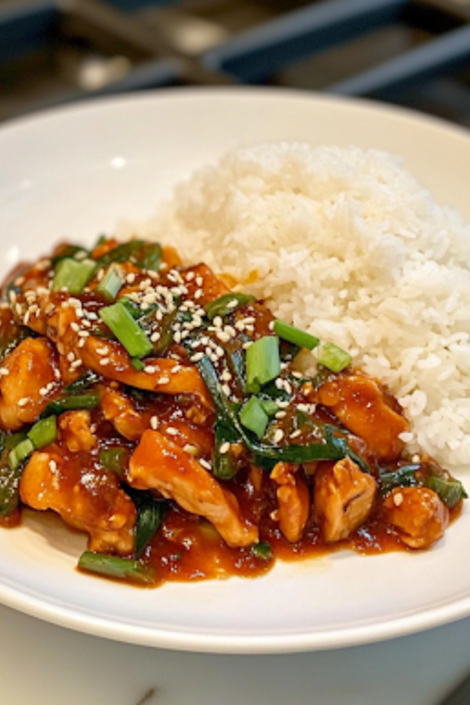 A close-up shot of a pair of chopsticks picking up a bite of perfectly glazed sweet chili chicken, garnished with sesame seeds and green onions. In the background, a bowl of steaming white rice sits on the white marble cooktop, complementing the delicious, saucy dish.