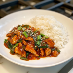 A close-up shot of a pair of chopsticks picking up a bite of perfectly glazed sweet chili chicken, garnished with sesame seeds and green onions. In the background, a bowl of steaming white rice sits on the white marble cooktop, complementing the delicious, saucy dish.