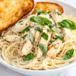A close-up shot of a serving plate on a white marble cooktop, featuring a generous portion of Chicken Alfredo Pasta, garnished with fresh basil and extra Parmesan. A side of golden garlic bread rests beside the plate, completing the comforting meal