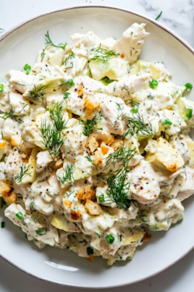 A close-up shot of a serving plate on a white marble cooktop, featuring the completed Avocado Chicken Salad. The dish is garnished with extra chives and dill, and crispy bread sits on the side, ready to be enjoyed.