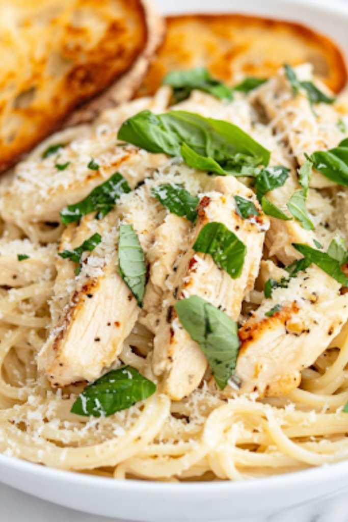A close-up shot of a serving plate on a white marble cooktop, featuring a generous portion of Chicken Alfredo Pasta, garnished with fresh basil and extra Parmesan. A side of golden garlic bread rests beside the plate, completing the comforting meal