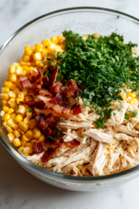 A close-up shot of the same glass bowl on a white marble cooktop, now filled with shredded chicken, sweet yellow corn kernels, crispy chopped bacon, finely chopped chives, and fresh dill. The ingredients create a vibrant medley of textures and colors.
