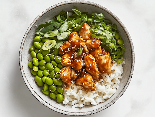 A close-up shot of the final plated Spicy Sesame Chicken Rice Bowl on a white marble cooktop, showcasing a vibrant dish of fluffy rice, crispy golden chicken, edamame, and fresh toppings, all ready to be enjoyed immediately.
