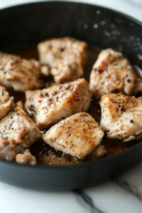 A close-up shot of the same black pot on a white marble cooktop, with bone-in chicken pieces lightly searing in a blend of whole peppercorns, a cinnamon stick, and aromatic spices. The surface of the chicken glistens as it absorbs the rich flavors