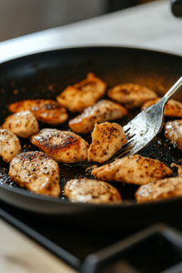 A close-up shot of a black skillet on a white marble cooktop, where seasoned chicken chunks sizzle and turn golden brown. The surface glistens with natural juices and a spatula stirs the pieces, ensuring even cooking