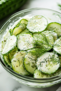 A bowl of chilled German cucumber salad served in a white ceramic dish on the white marble cooktop. The creamy dressing lightly coats the thinly sliced cucumbers, with fresh dill sprinkled on top as a garnish, ready to be enjoyed.