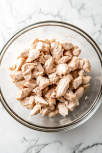 A close-up shot of a glass mixing bowl placed on a white marble cooktop, filled with bite-sized pieces of chopped cooked chicken, ready to be mixed with other ingredients.