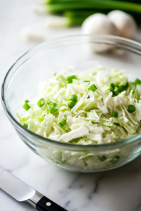 A glass mixing bowl on a white marble cooktop holds finely chopped cabbage and sliced green onions, neatly arranged. A sharp knife rests beside the bowl, ready for more ingredient preparation.