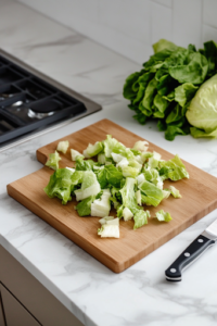 A close-up view of a wooden cutting board on a white marble cooktop, where freshly washed romaine lettuce is being chopped into bite-sized pieces. A sharp knife rests beside the board, with whole romaine leaves nearby, ready for preparation.