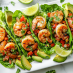 A close-up shot of fully assembled garlic cilantro lime shrimp lettuce wraps arranged on a white serving platter over a white marble cooktop. Lime wedges, avocado slices, and diced tomatoes are placed around the wraps, making them look fresh and ready to be served.