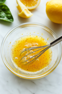 A close-up shot of a small glass bowl on the white marble cooktop, containing a freshly mixed blend of orange juice, lemon juice, lemon zest, and a pinch of salt. A whisk is actively stirring the mixture, blending the tangy and aromatic citrus dressing for the Summer Citrus Fruit Salad.