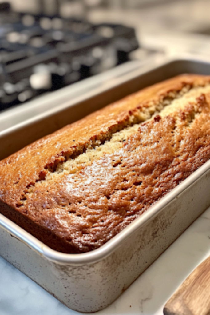 A close-up of a freshly baked golden-brown banana bread loaf cooling in its pan on a white marble cooktop. A knife rests beside the pan, ready to slice into the moist, fluffy bread, revealing its soft and tender crumb.