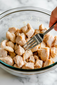 Overhead shot of a glass mixing bowl with diced chicken pieces marinating in a dark, glossy mixture of soy sauce, chili vinegar, cornstarch, and ground ginger. A spoon stirs the ingredients evenly.