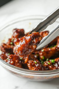 A close-up shot of a glass bowl on a white marble cooktop, where the seasoned chicken drumsticks are being coated in a rich, golden glaze. A pair of tongs lifts a drumstick, its surface glistening with the sticky, flavorful sauce, ensuring an even coating for maximum taste.