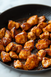 A black skillet on a white marble cooktop is filled with golden-brown chicken chunks coated in the thick, caramelized honey bourbon glaze. The sauce clings to each piece, creating a glossy and irresistible finish.