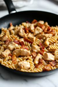 A close-up shot of the black skillet on a white marble cooktop, as cooked rotini pasta, golden seared chicken, and crispy crumbled bacon are tossed together in the luscious creamy ranch sauce. Every piece is coated evenly, ensuring a perfectly combined dish.