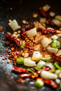 Close-up of a wok with sliced garlic, minced ginger, dried red chilies, Sichuan peppercorn powder, and scallions sizzling in hot oil. The vibrant colors and aromatic ingredients are in focus.
