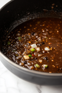 Inside the black pot on the white marble cooktop, softened onions, bell peppers, celery, and garlic are being stirred into the dark roux. The vegetables glisten as they cook down, releasing their fragrant aroma into the kitchen.