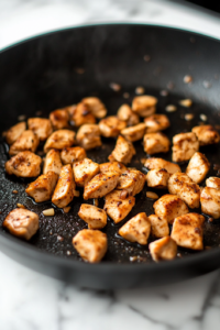 A close-up shot of the same black skillet on the white marble cooktop, now with cubed chicken pieces added. The chicken sears to a golden brown, soaking up the flavors of garlic and spices, while the air fills with a mouthwatering aroma.